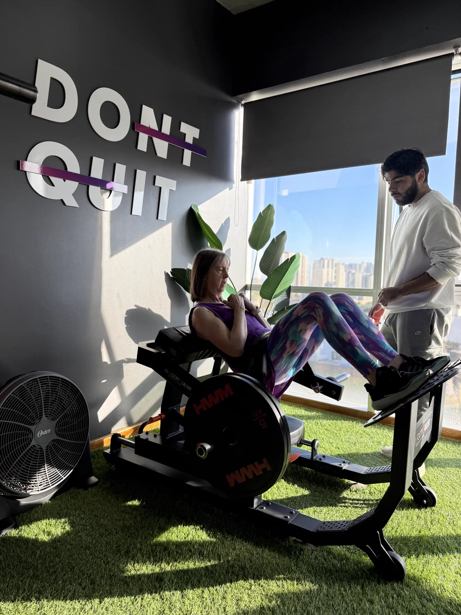 Entrenador acompañando a una clienta en máquina de empuje de cadera, junto al muro DON'T QUIT del estudio.